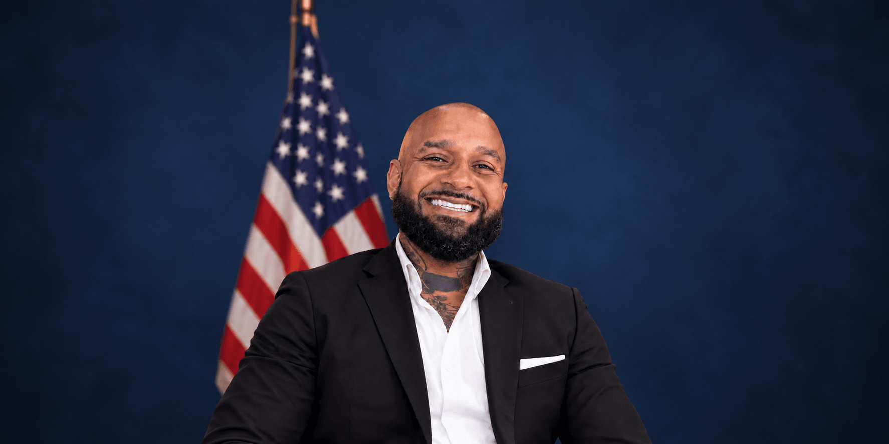 Smiling man in a black suit seated against an American flag background