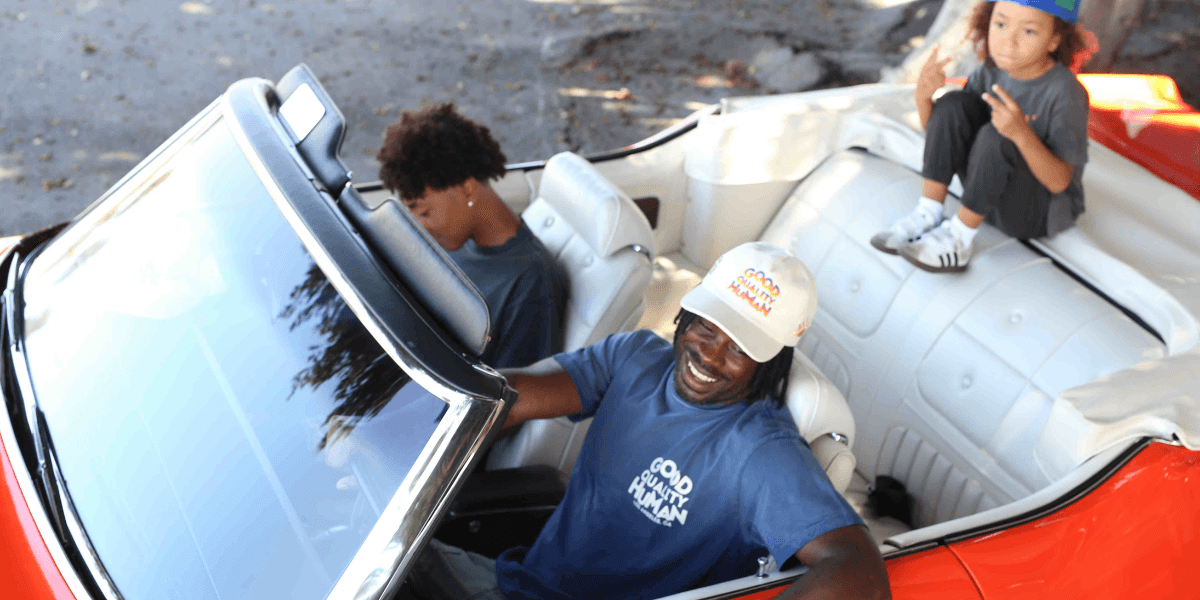 A smiling man drives a classic convertible with two kids inside.