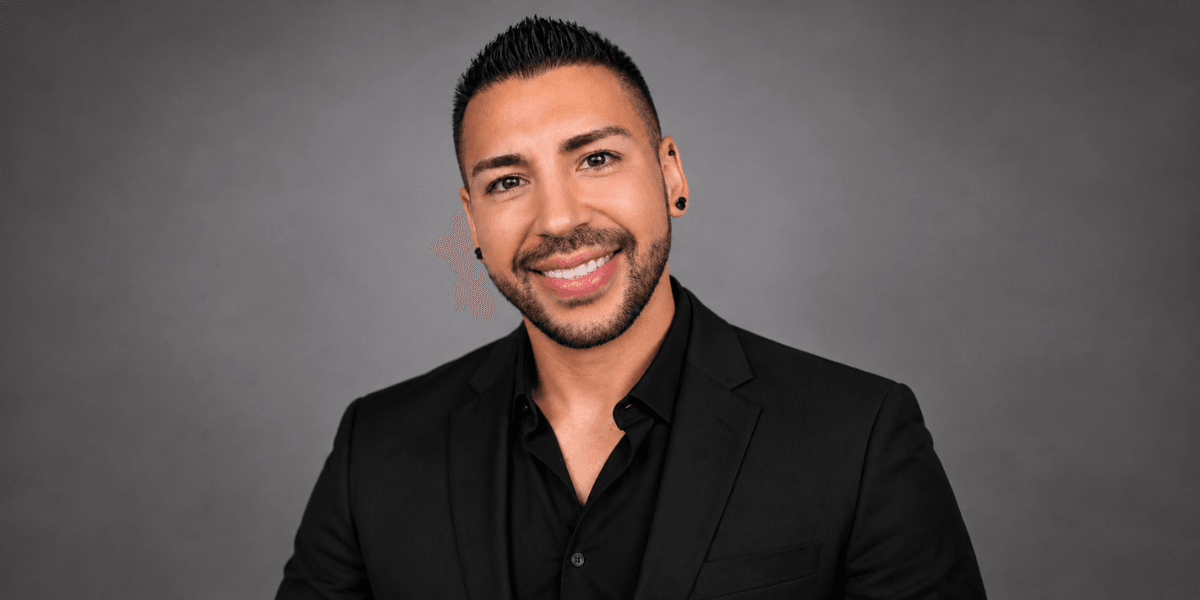 Man in a black suit shirt smiling against a grey backdrop