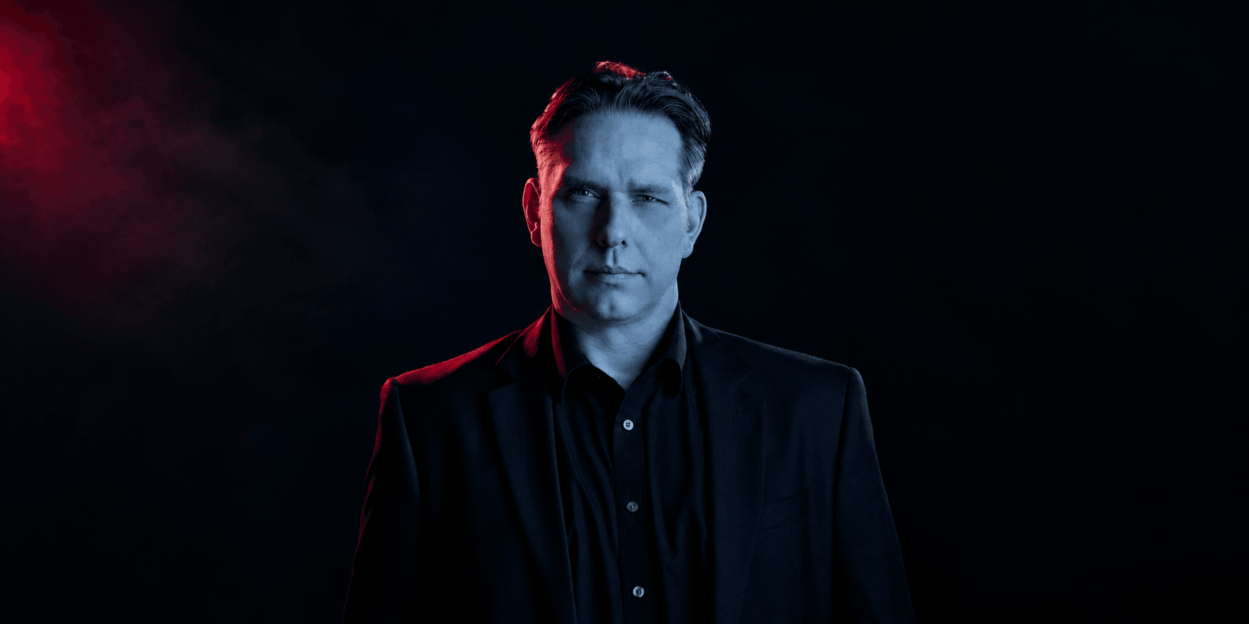 A man in a dark suit stands against a smoky, dramatic background.