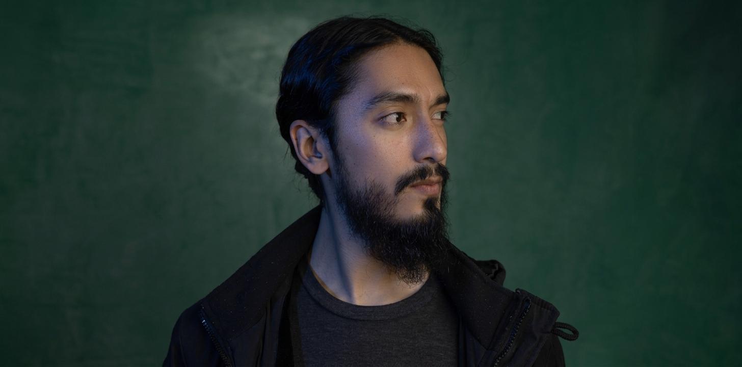 A man with long black hair and a beard poses against a green background