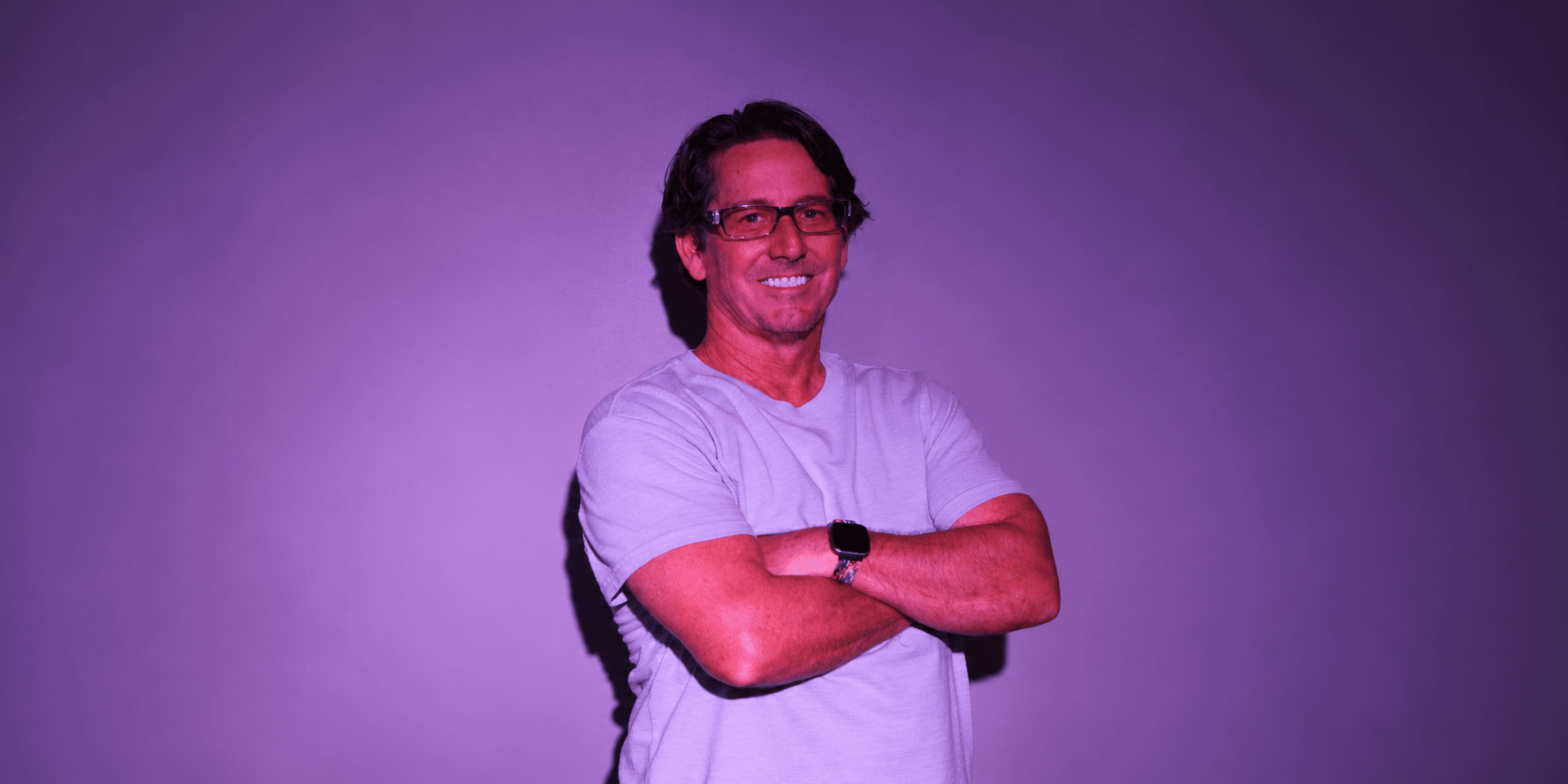 Smiling man in a light gray shirt stands with arms crossed against a purple backdrop