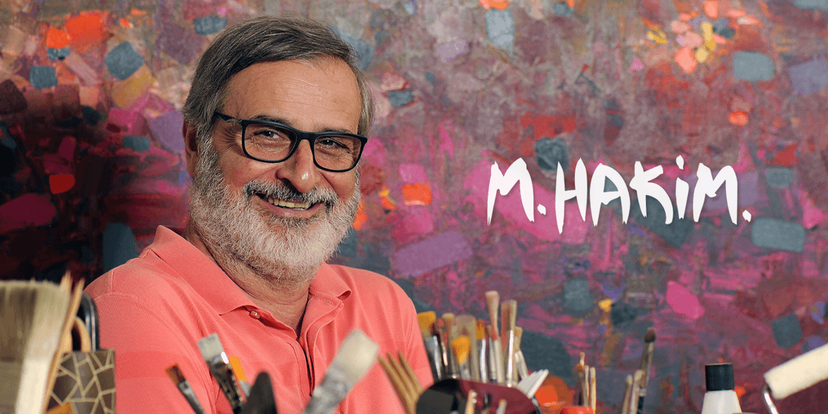 "A smiling artist, M. Hakim, wearing a pink polo shirt and glasses, standing in front of a vibrant abstract painting with art tools surrounding him."