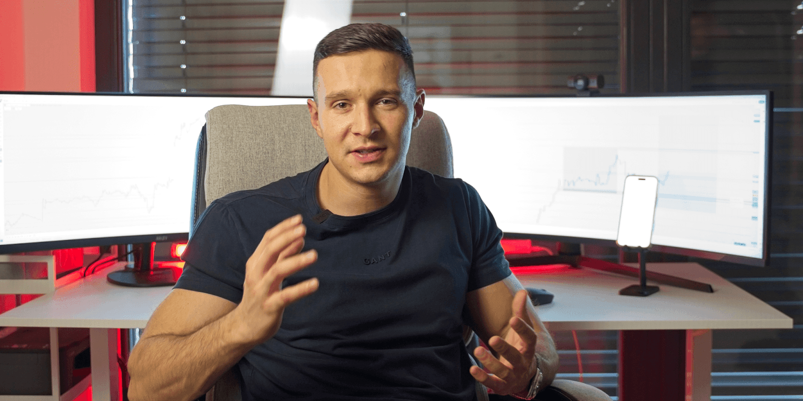 Michal, a top-performing forex trader in Central Europe, sits in front of dual trading monitors displaying live forex charts. He gestures confidently while discussing market strategies, wearing a dark shirt with a headset mic attached. The trading setup behind him includes a smartphone on a stand and red ambient lighting, emphasizing a high-performance, professional environment.