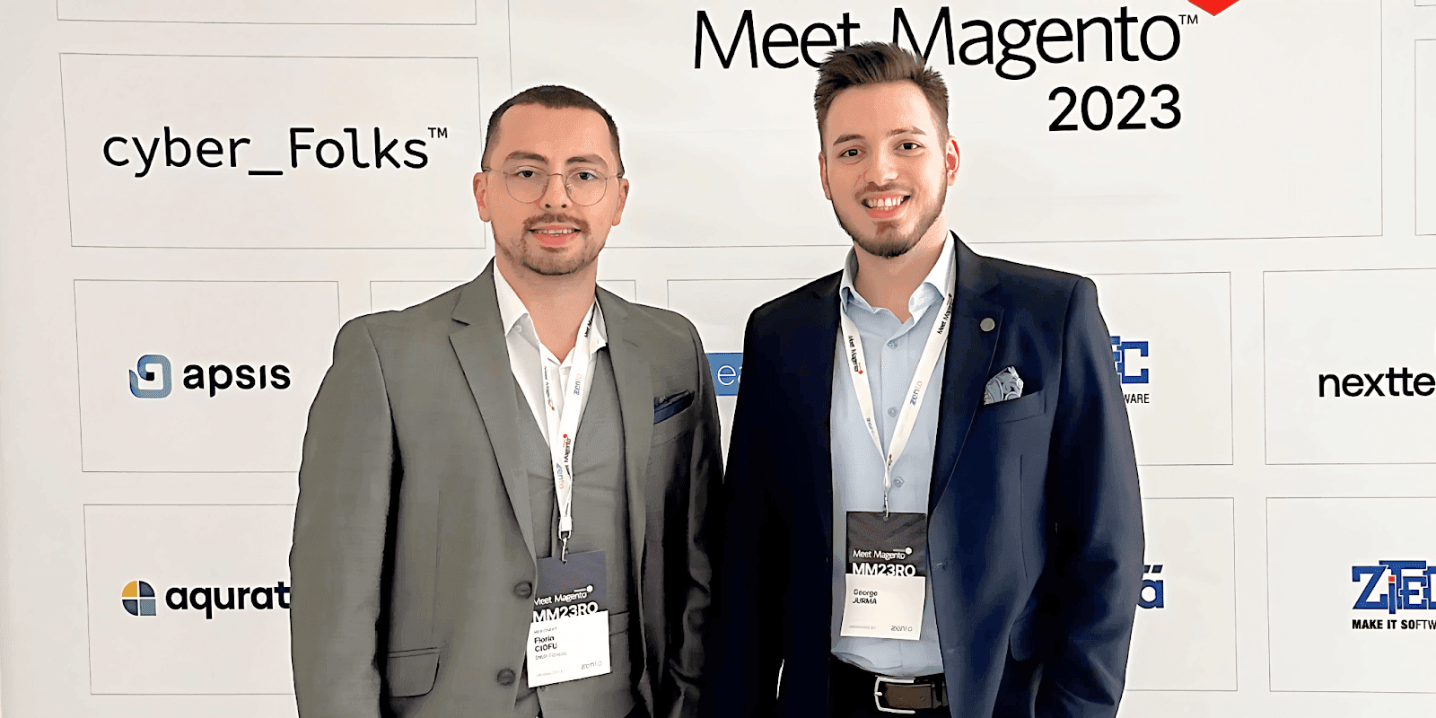 Two men standing together in business attire at a Meet Magento event, with a backdrop of event logos.