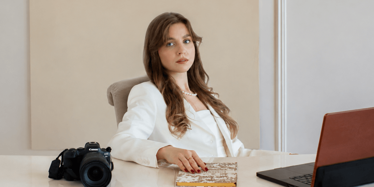 A woman in a white suit seated at a desk with a camera and laptop