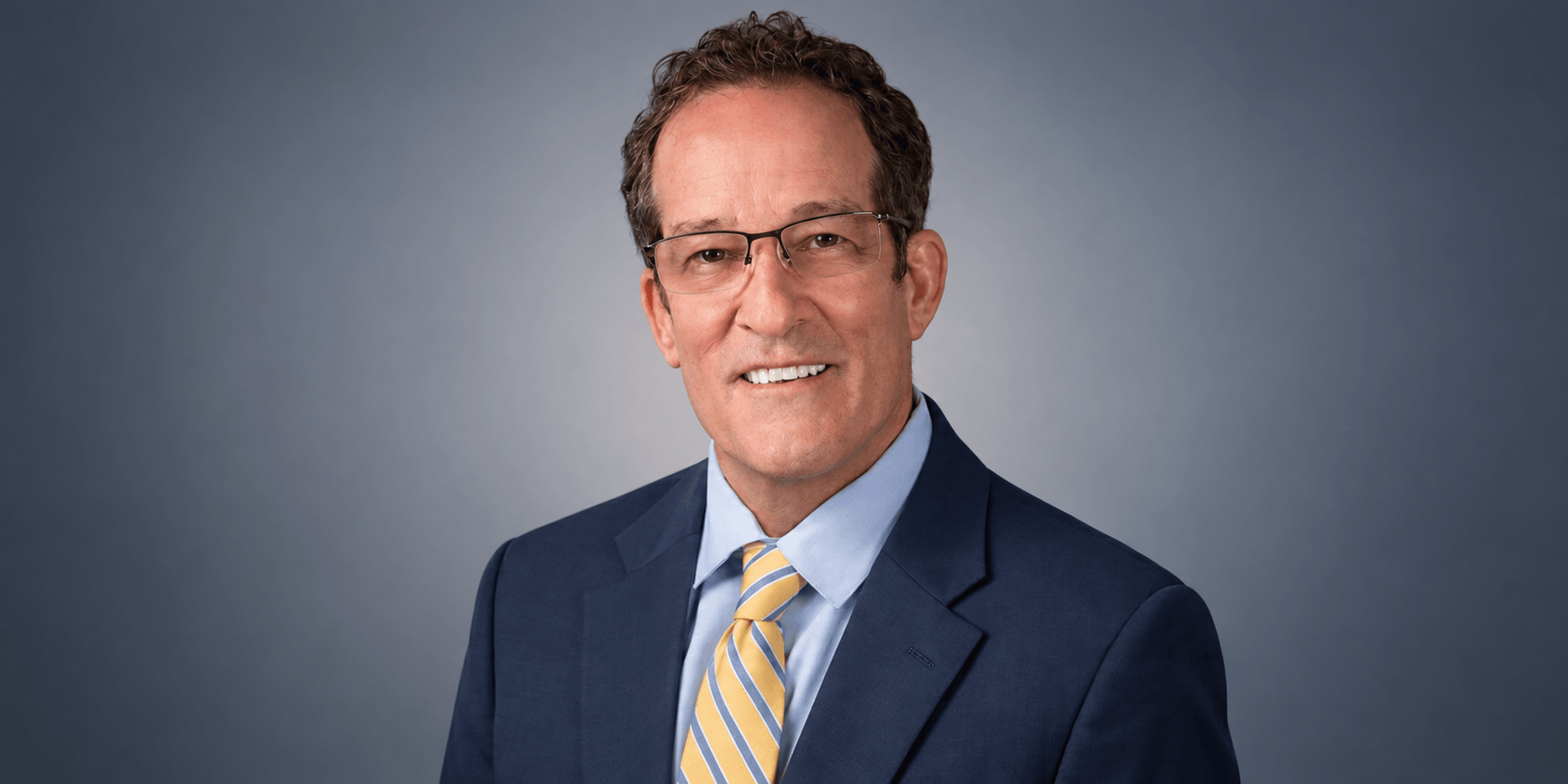A middle-aged man in a suit and glasses smiling against a gray background