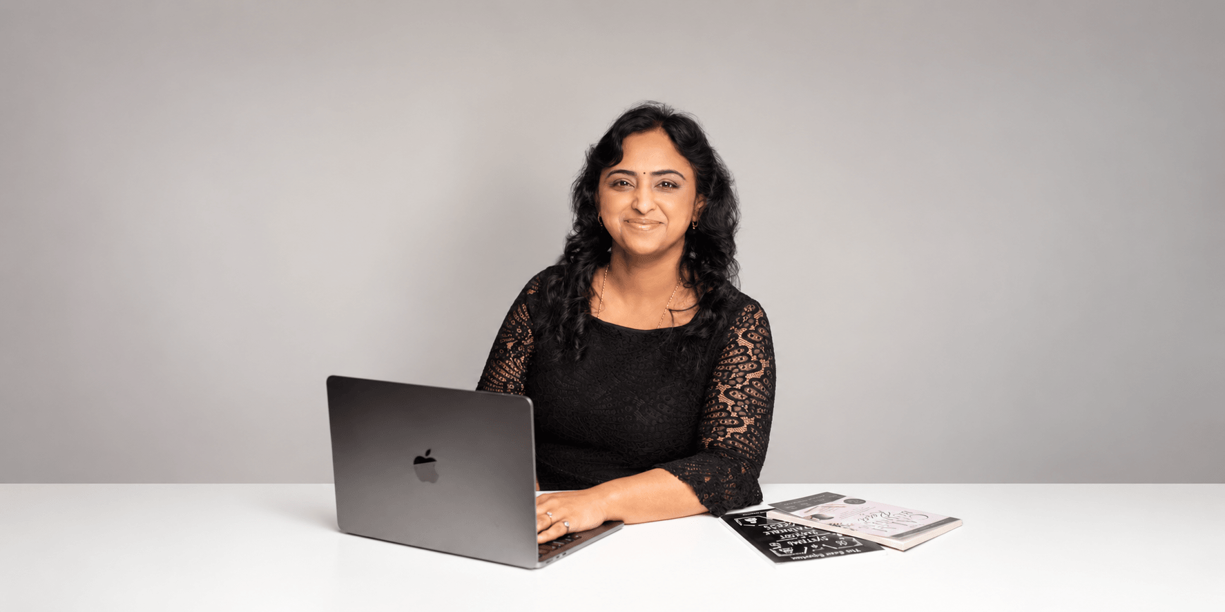 A woman with black curly hair sits at a desk with a laptop and notebooks