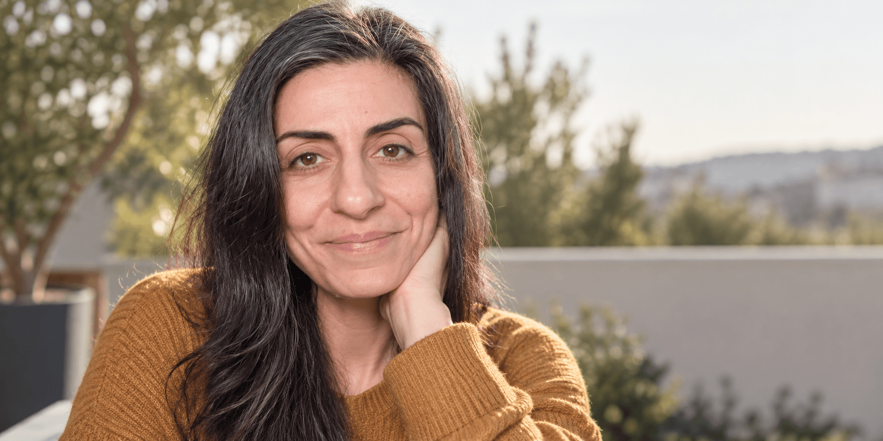 A woman with long dark hair smiles, resting her chin on her hand outdoors.