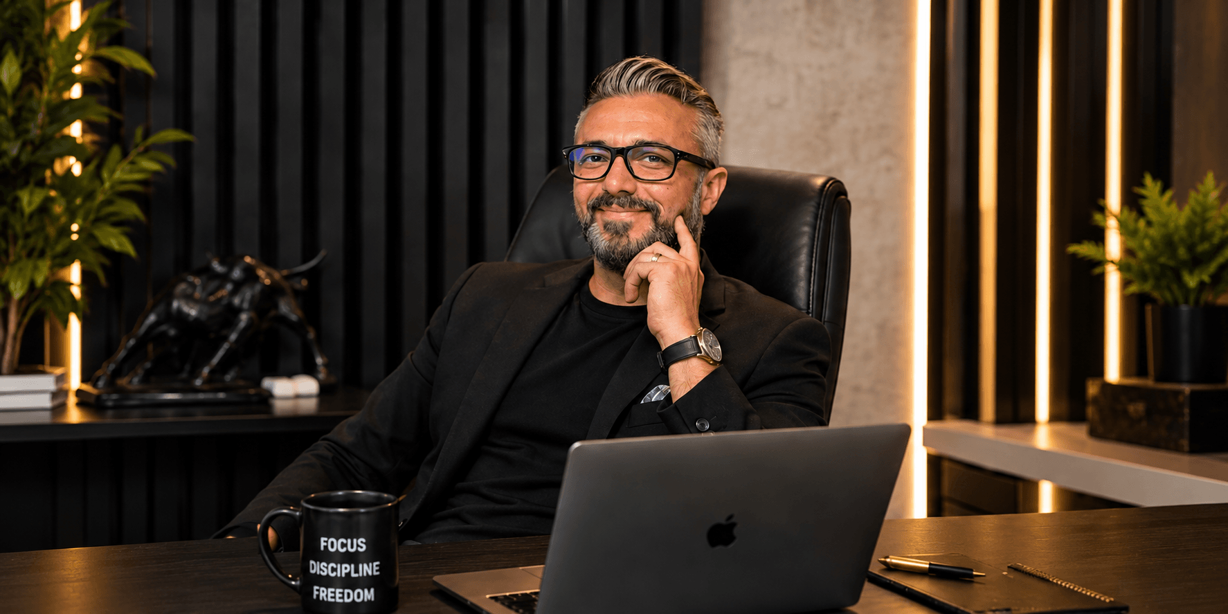 Man in a black suit sitting at a desk, smiling with a laptop and coffee mug