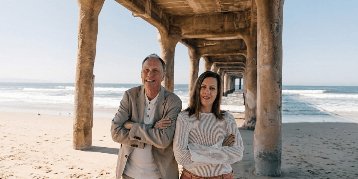 Two people smile under a pier by a beach, with waves in the background