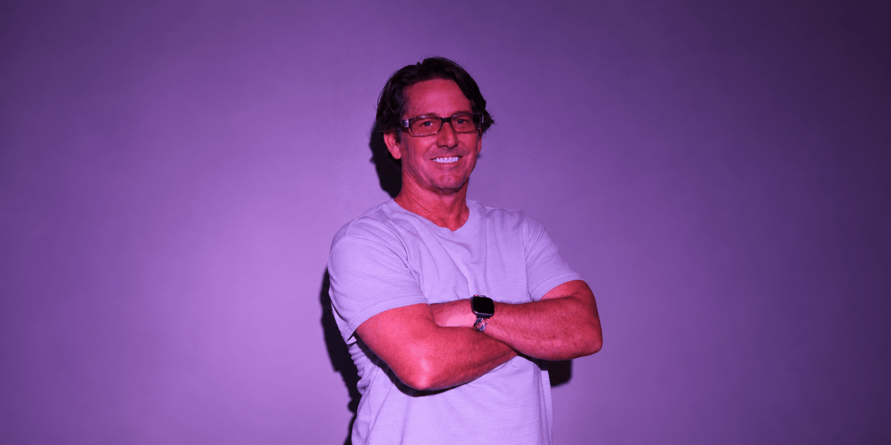 Smiling man in a light gray shirt stands with arms crossed against a purple backdrop