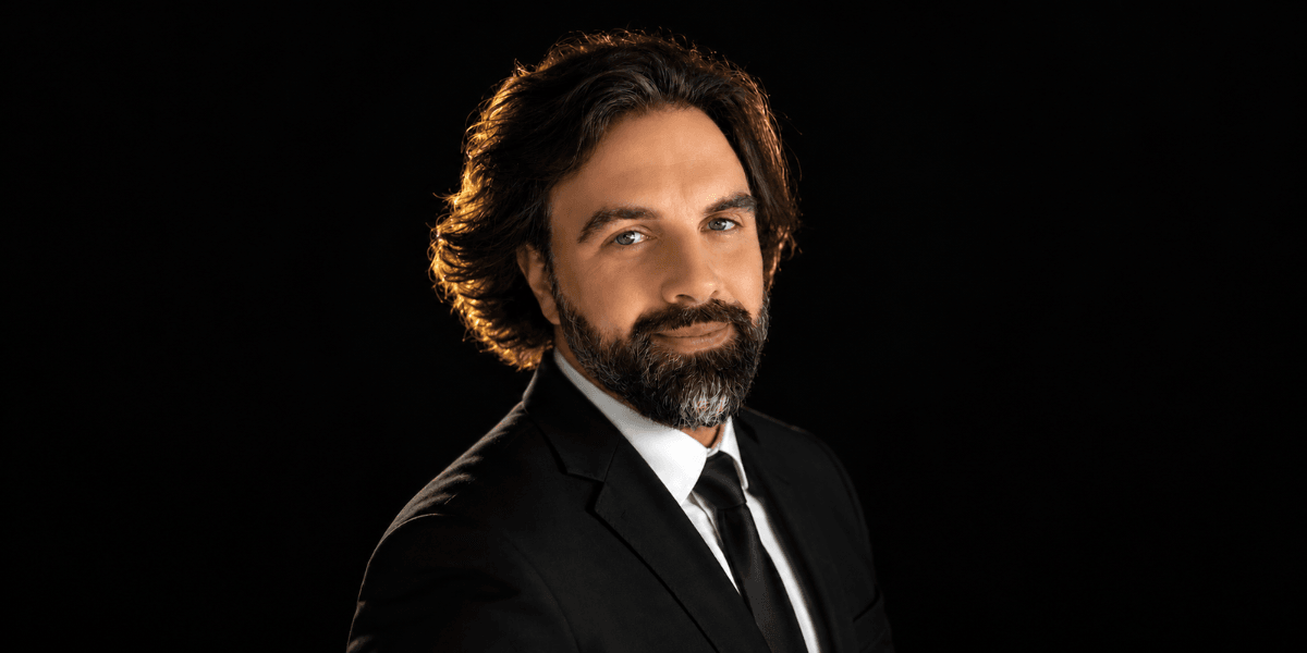 Man in a suit with a beard poses confidently against a black background