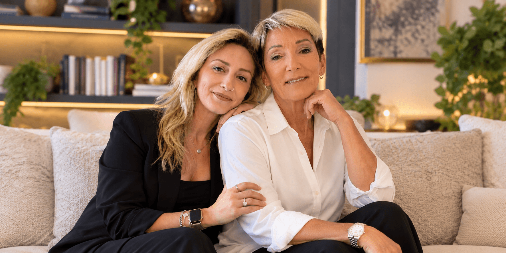 Two women sitting on a sofa, smiling, with plants and books in the background