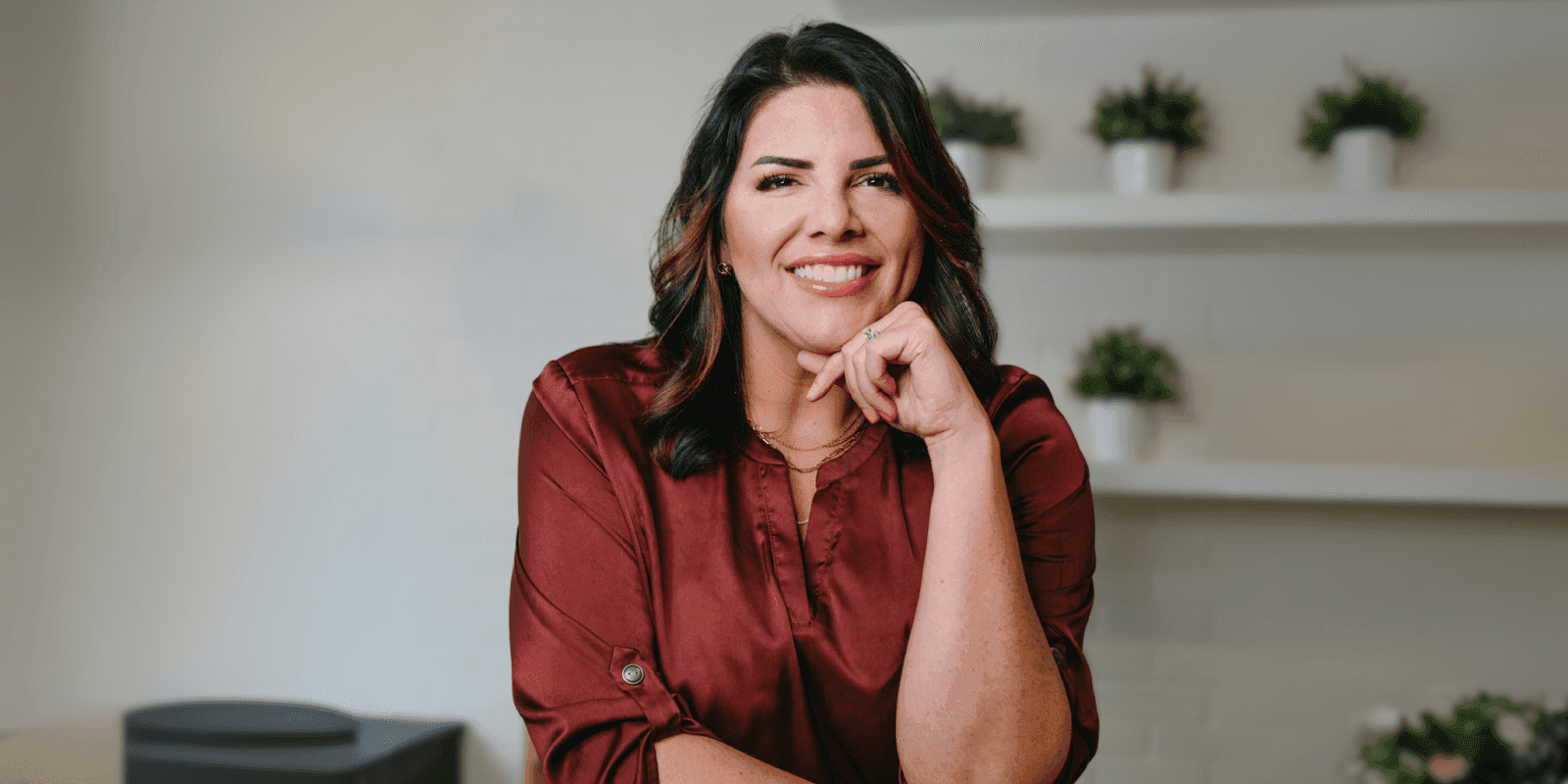 Smiling woman in maroon blouse poses indoors with plants on shelves behind her.