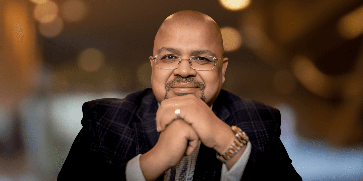 Man in glasses and suit jacket smiling with hands clasped under chin.