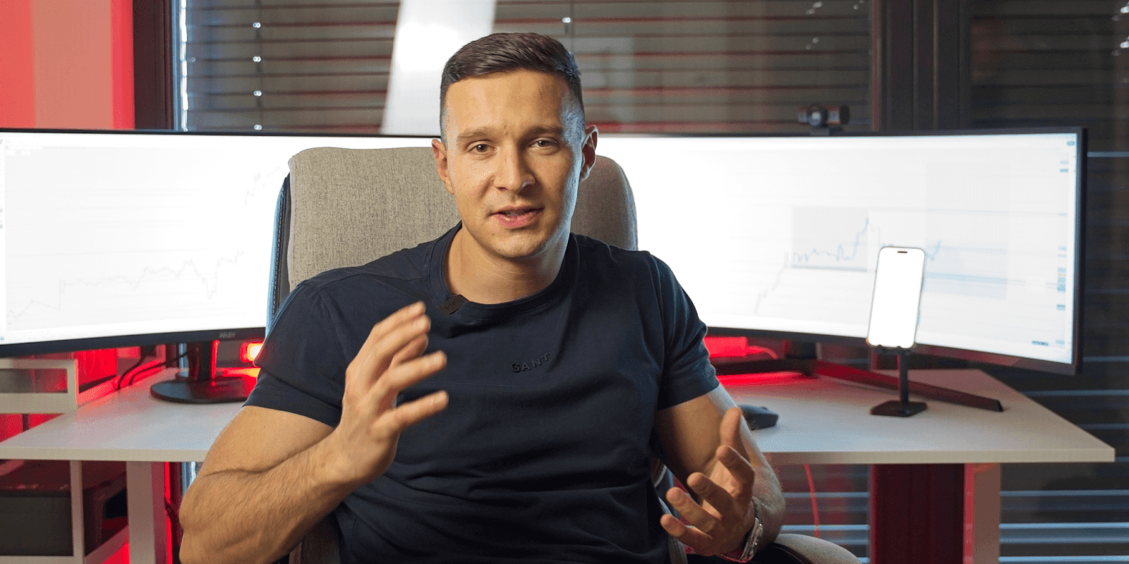 Michal, a top-performing forex trader in Central Europe, sits in front of dual trading monitors displaying live forex charts. He gestures confidently while discussing market strategies, wearing a dark shirt with a headset mic attached. The trading setup behind him includes a smartphone on a stand and red ambient lighting, emphasizing a high-performance, professional environment.