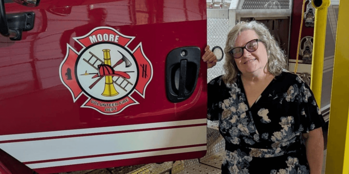 Mary Myers standing beside a Moore Volunteer Fire Dept truck, smiling and wearing glasses and a floral dress.