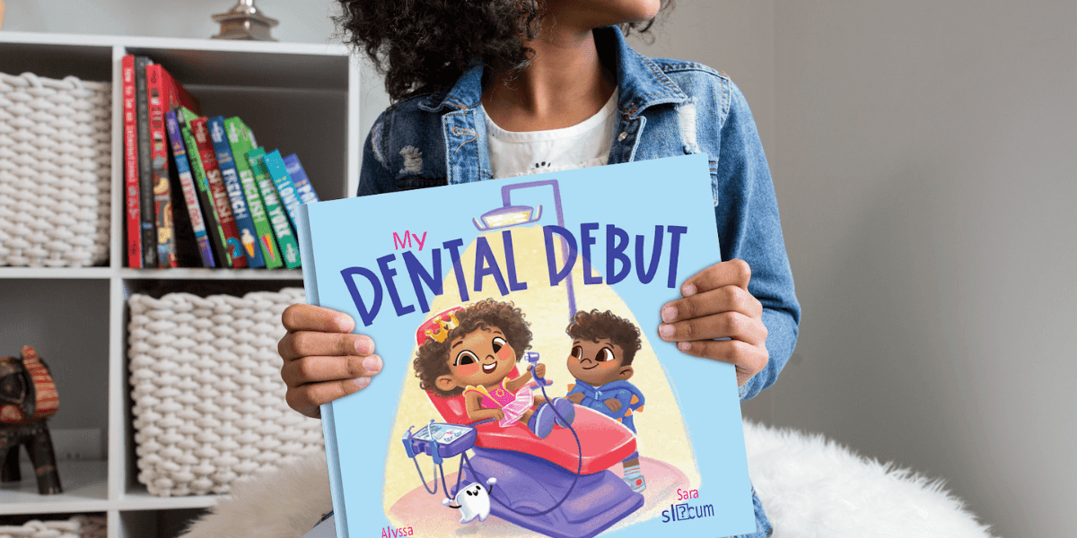 A young African American girl is shown holding a children's book titled 'My Dental Debut' in a cozy room, her curly hair framing her face as she gazes to the right. The image captures vibrant colors, from the book’s dreamy blue cover featuring a cheerful dental scene to the vivid spines of children's books on a nearby shelf, all bathed in soft, natural light with a welcoming atmosphere.