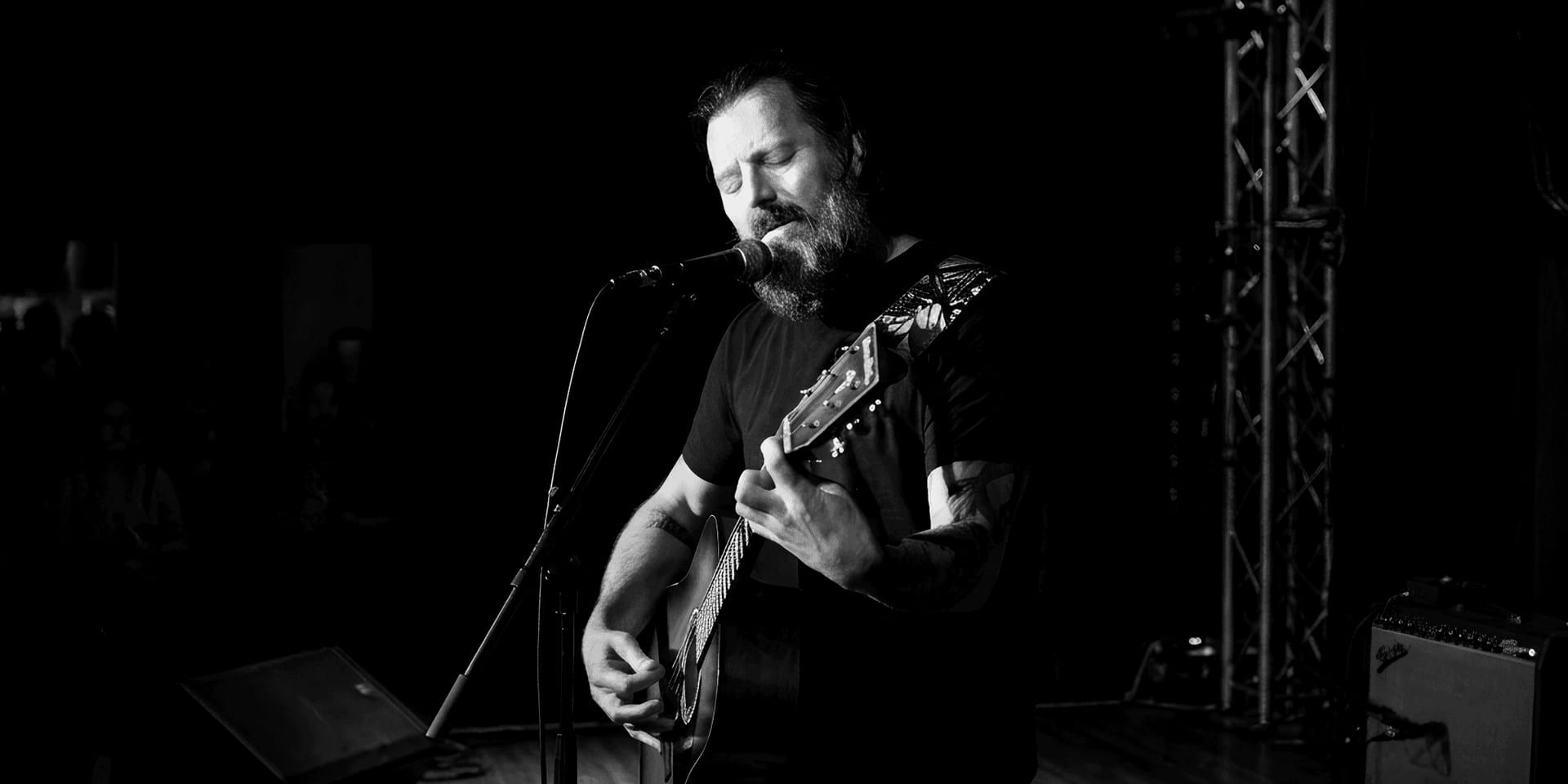Scott Lennox sings and plays acoustic guitar during an intimate live performance under stage lighting.