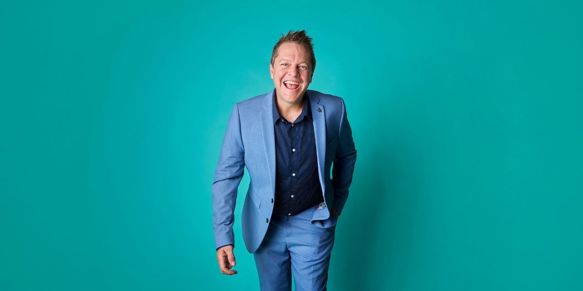 A professional man wearing a blue suit, smiling in front of a bright turquoise background.