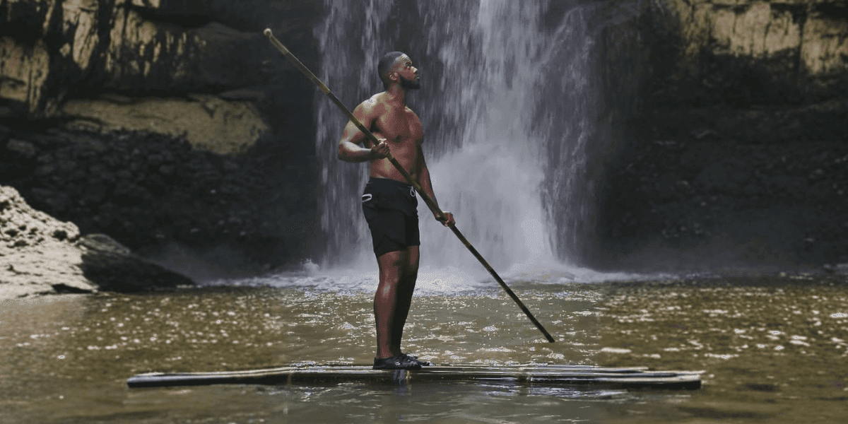 Chaz Ferguson paddleboards in front of a waterfall, holding a paddle in calm water.