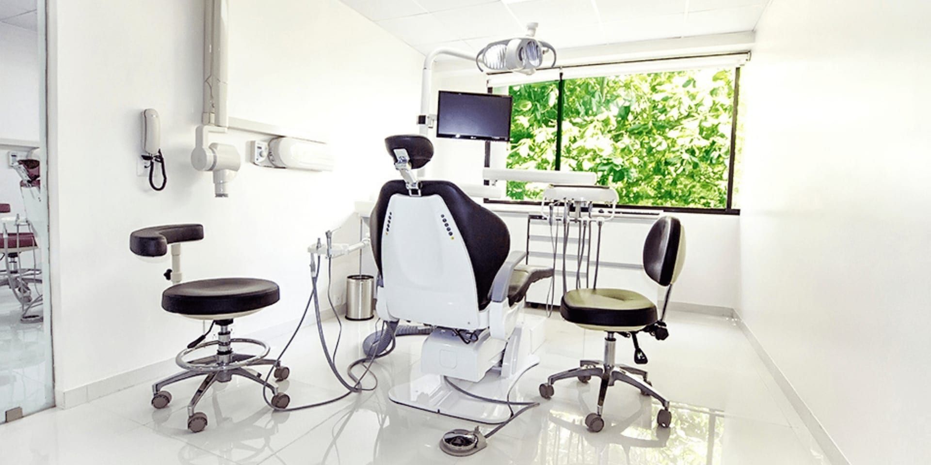 Modern and clean dental clinic interior featuring a dentist chair, a monitor mounted on the wall, and two ergonomic chairs for patients, with natural light streaming in through a large window showcasing a lush green view.