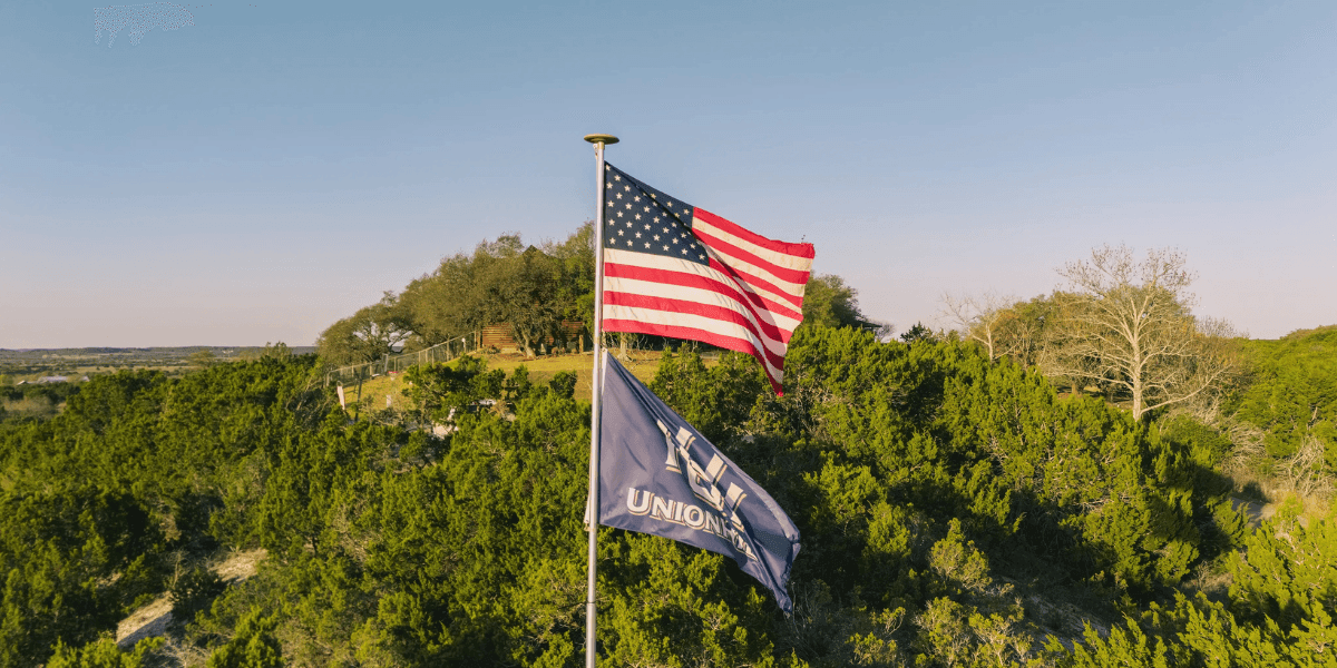 American and Union Hill Ranches flags flying over Texas Hill Country landscape near a luxury hunting and hospitality lodge.