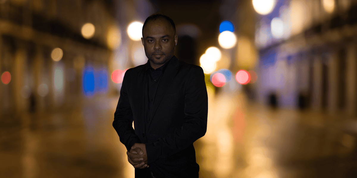 "A confident man in a black suit standing in a dimly lit street at night, with colorful bokeh lights in the background, exuding a professional and polished aura."