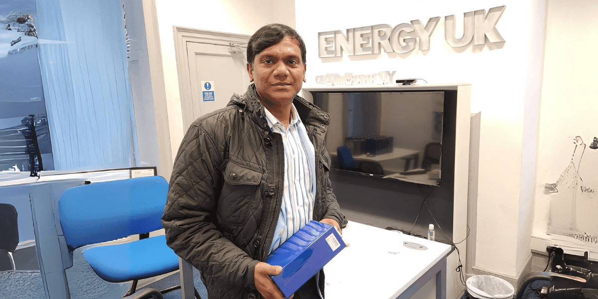Man holding a blue box in front of the ENERGY UK sign and a TV screen in an office setting.