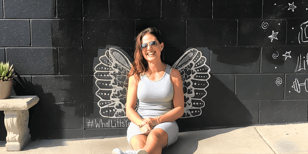 Angela Porrelli sitting in front of a mural with illustrated wings, smiling outdoors in sunglasses.