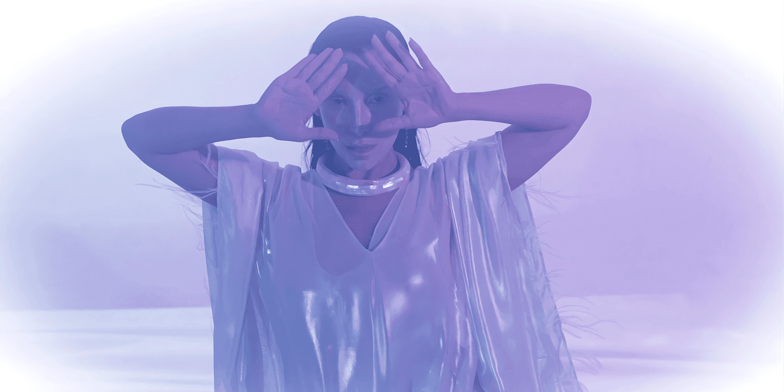 A woman in a metallic silver outfit with a glowing choker, performing with a mystical, ethereal aura in purple light.