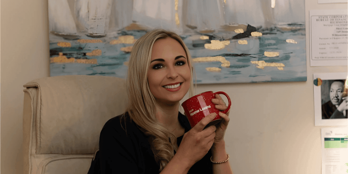 Sheri Wall, a dedicated loan officer at Keller Home Loans, smiles while holding a red mug with the logo for "Home Loans." She is seated in an office with a background featuring a serene painting of water and gold accents. Sheri's warm and approachable demeanor reflects her commitment to guiding clients through the home buying process with clarity and care, especially military families and first-time homebuyers. Ask ChatGPT