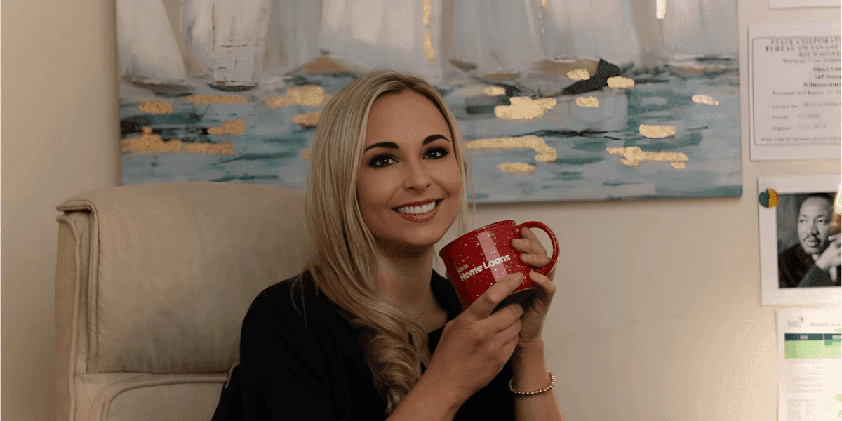 Sheri Wall, a dedicated loan officer at Keller Home Loans, smiles while holding a red mug with the logo for "Home Loans." She is seated in an office with a background featuring a serene painting of water and gold accents. Sheri's warm and approachable demeanor reflects her commitment to guiding clients through the home buying process with clarity and care, especially military families and first-time homebuyers. Ask ChatGPT