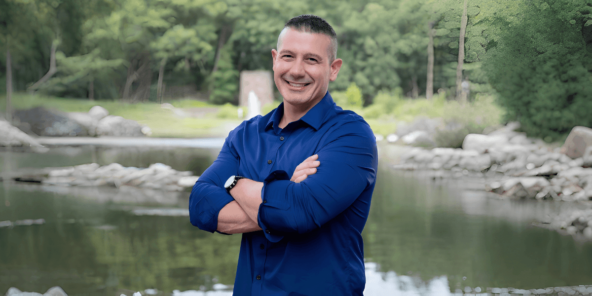 Bill Yeager smiles with folded arms beside a peaceful river, representing leadership, fitness, and personal transformation.