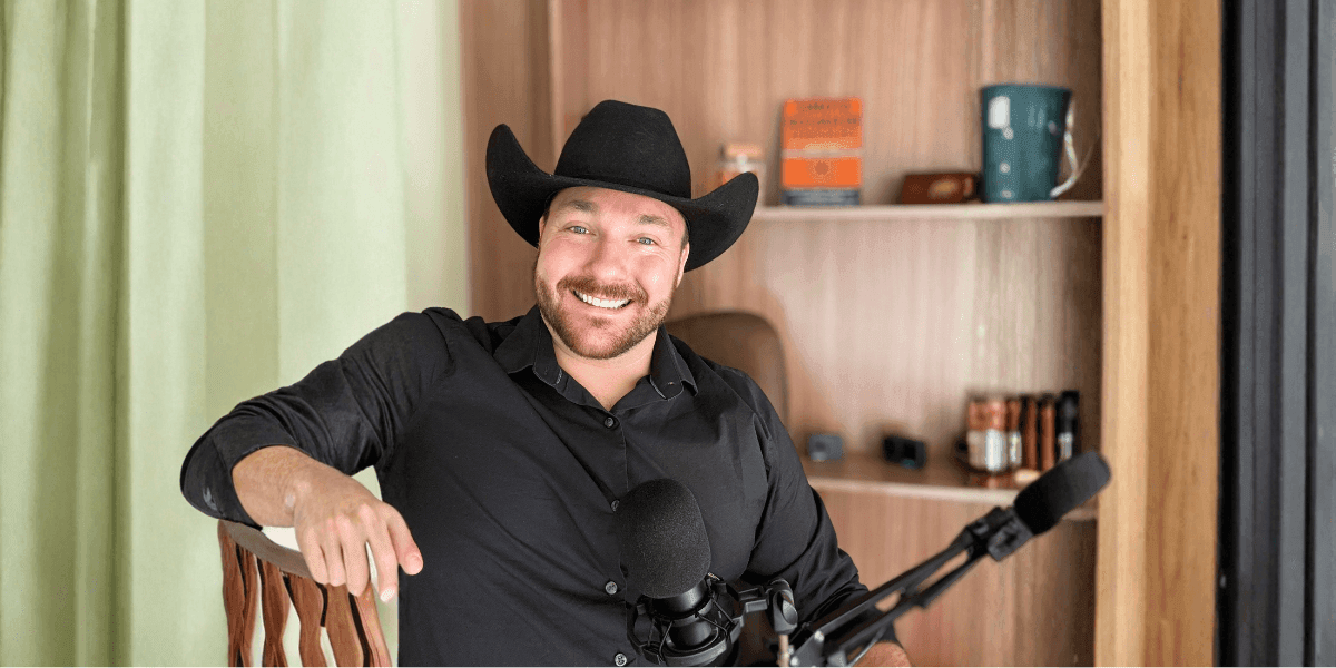 a man wearing a cowboy hat, smiling while sitting beside a microphone in a podcast setup.