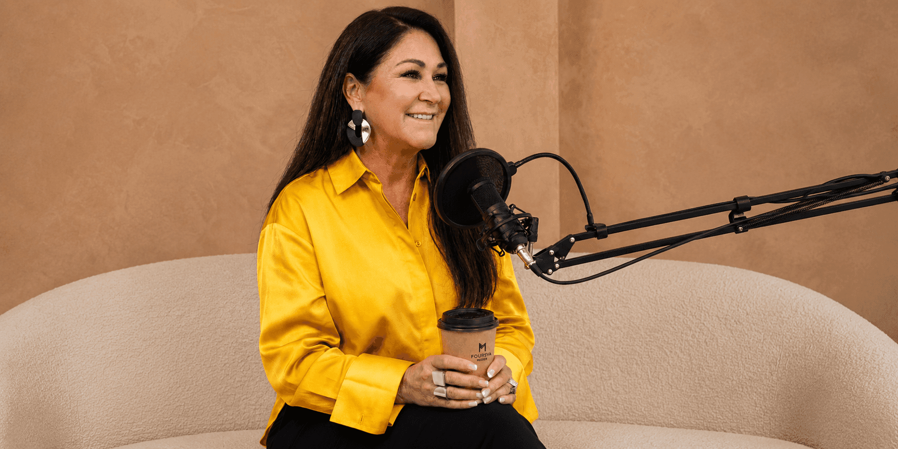 Traycee Mayer, a woman on a couch, holds a microphone and coffee, symbolizing her focus on connection in leadership.