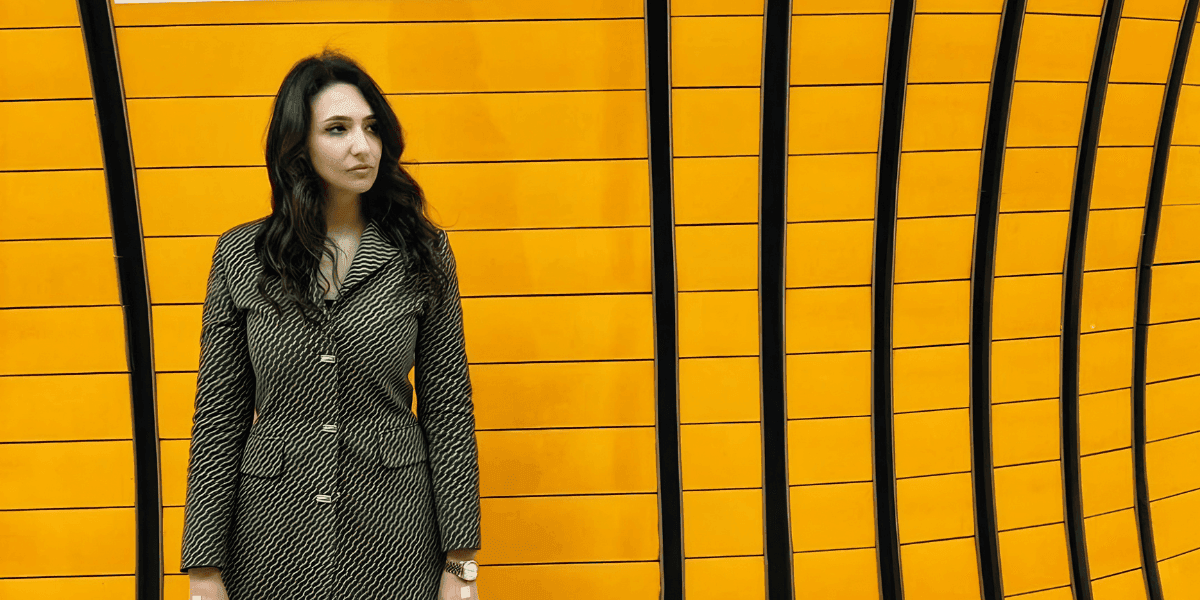 A woman wearing a green coat poses in front of a vibrant yellow wall, representing innovation in SaaS and education technology.