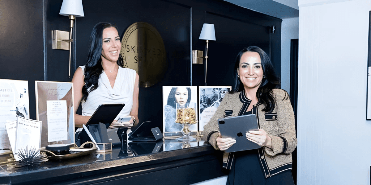 Inside Skinmed Spa with two women at the front desk, one holding an iPad and the other assisting customers with a smile.