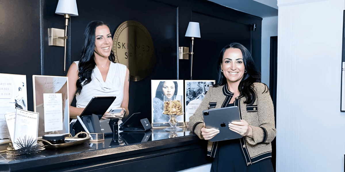 Inside Skinmed Spa with two women at the front desk, one holding an iPad and the other assisting customers with a smile.