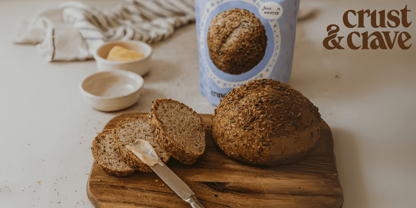 "Sliced loaf of Crust & Crave bread with butter spread on one slice, placed on a wooden cutting board, with the bread mix packaging in the background."