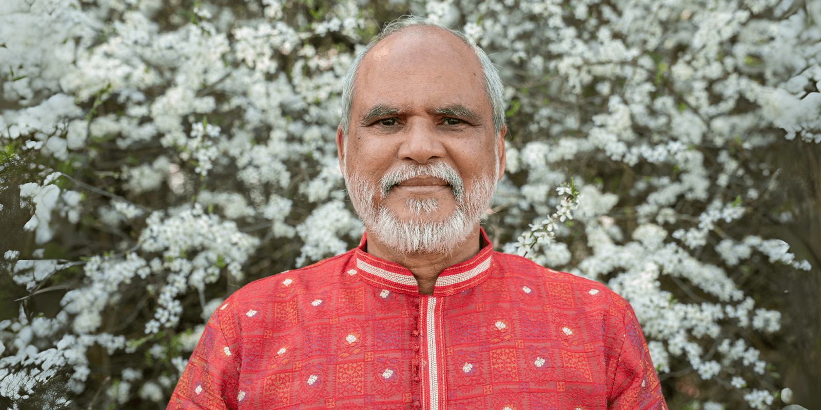 Smiling elderly man dressed in a red traditional outfit standing in front of a flowering tree, radiating positivity.