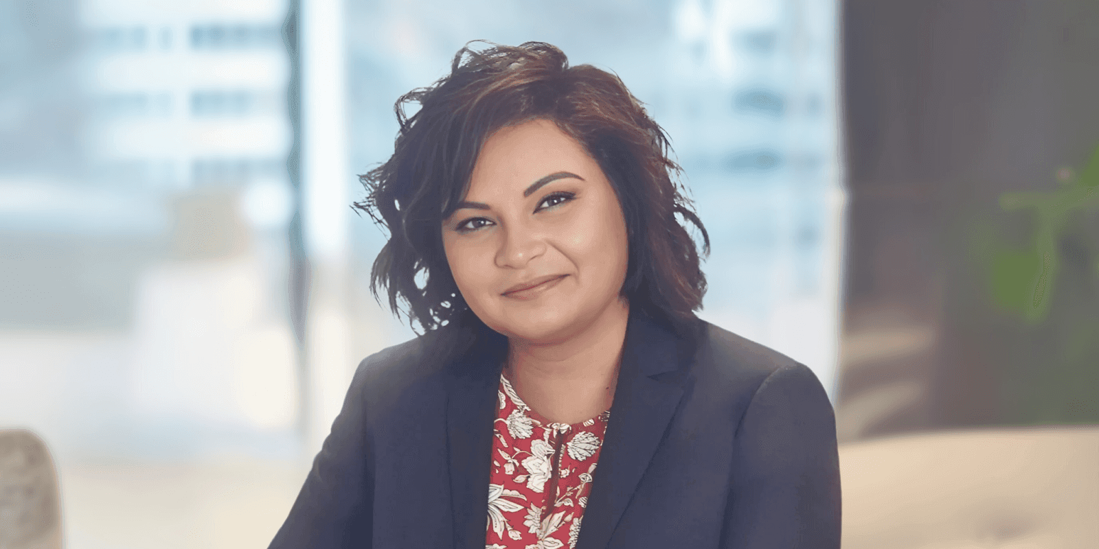 "Professional woman with short hair, wearing a blazer, smiling confidently in a business environment with a modern city backdrop."