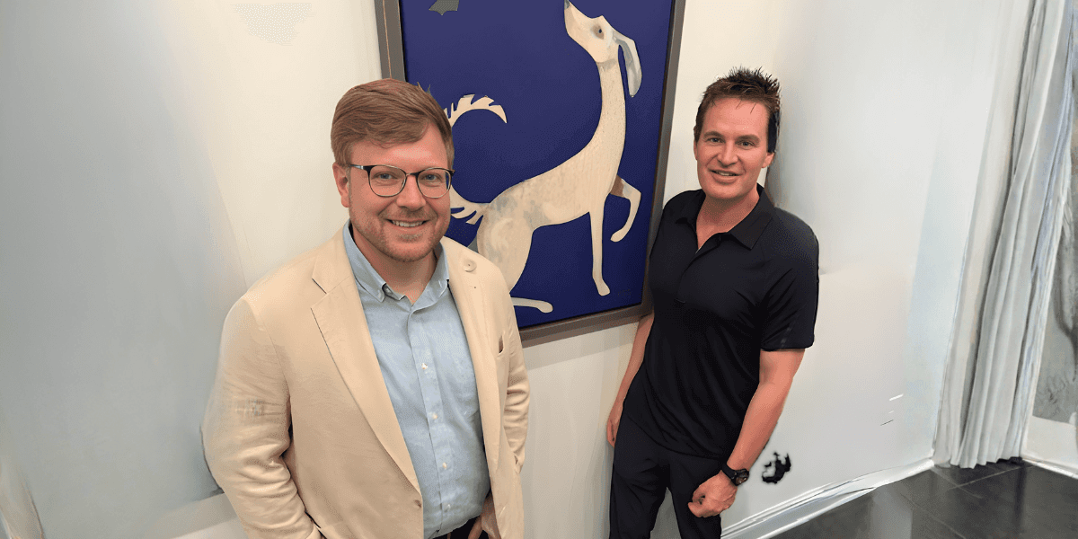 Brett Campbell and another man stand beside artwork of a white dog, representing his role connecting artists and collectors.