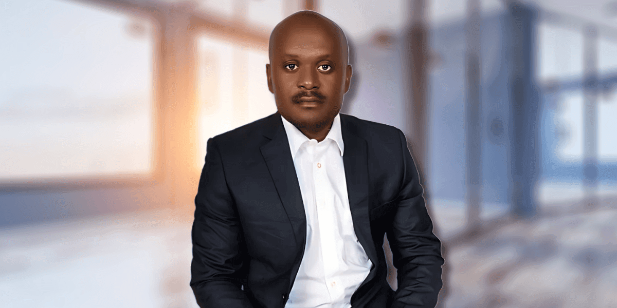 "Professional portrait of a man in a dark suit and white shirt, sitting confidently in a modern office setting with a blurred background."