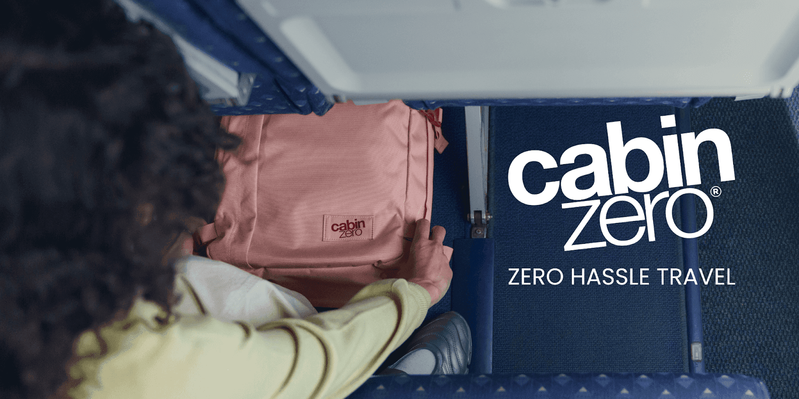 Person placing a pink CabinZero backpack under airplane seat with “Zero Hassle Travel” slogan displayed.