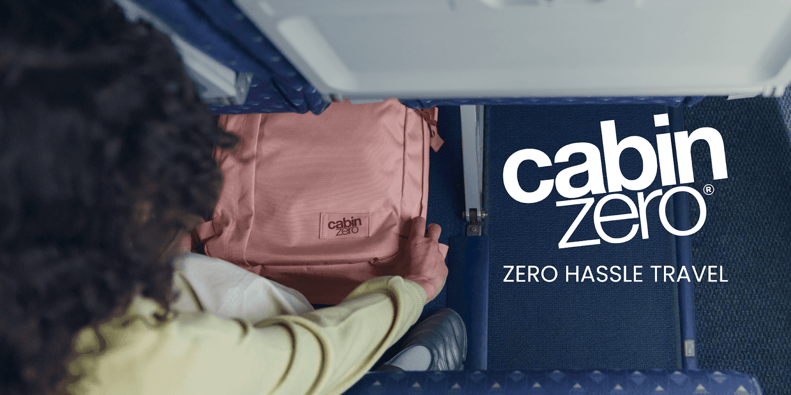Person placing a pink CabinZero backpack under airplane seat with “Zero Hassle Travel” slogan displayed.