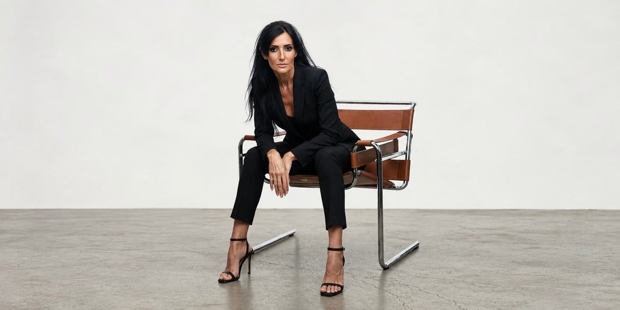 Woman in a black suit sitting on a modern chair against a neutral backdrop