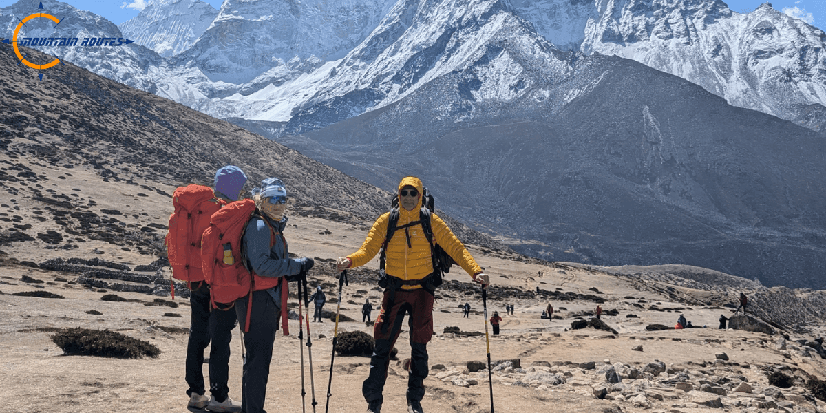 Three hikers with trekking poles and large backpacks standing in a rocky alpine landscape with snow-capped mountains in the background, part of a guided expedition with Mountain Routes.