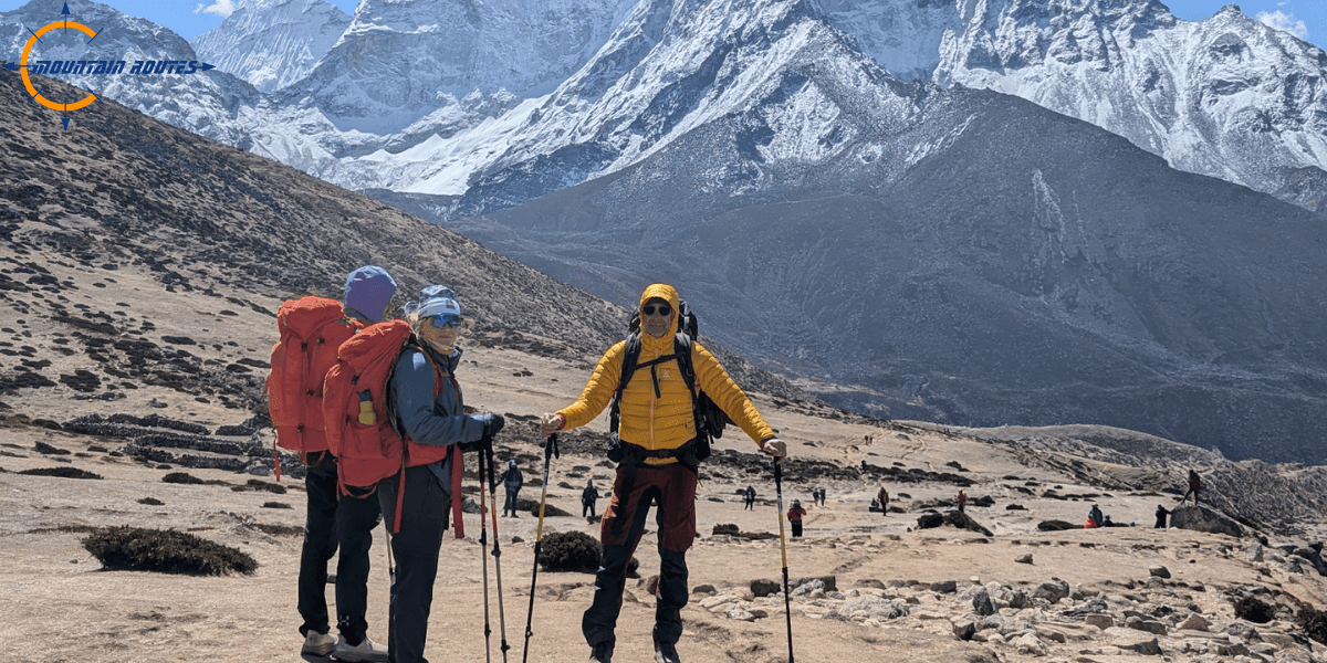 Three hikers with trekking poles and large backpacks standing in a rocky alpine landscape with snow-capped mountains in the background, part of a guided expedition with Mountain Routes.
